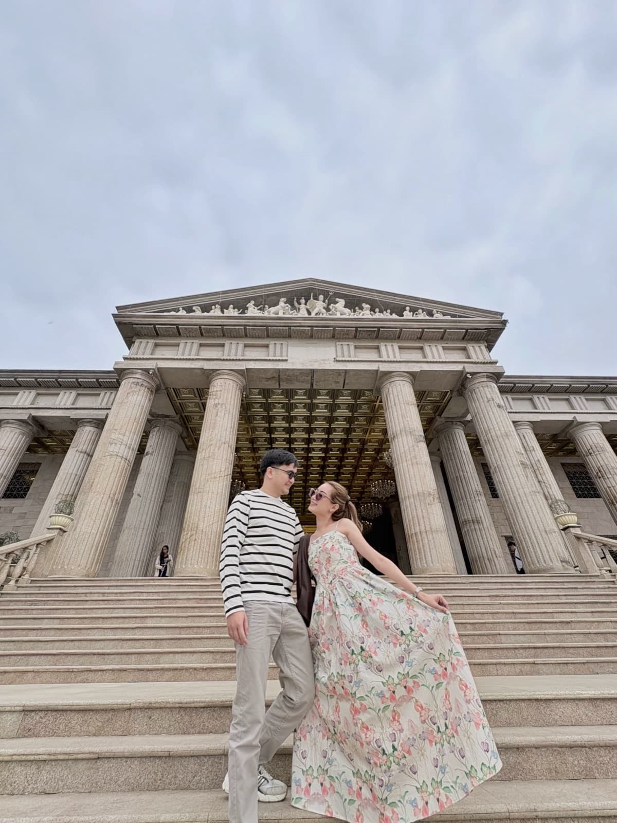Justin and Lovely smiling at each other on the steps of a grand columned building, Lovely in a floral gown caught mid-twirl