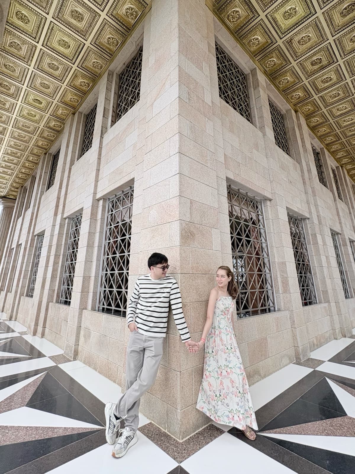 Justin and Lovely leaning against a stone column under a gilded ceiling, holding hands on a black and white geometric-tiled floor